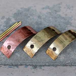 May include: Three metal strips in different finishes: copper, brass, and polished brass. Each strip is curved and has two holes. The strips are arranged in a row, with the copper strip on the left, the brass strip in the middle, and the polished brass strip on the right.