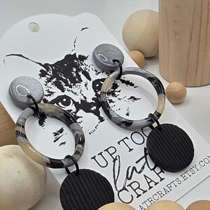 May include: A pair of handmade earrings with a gray and black marbled design. The earrings have a circular hoop and a textured black disc. The earrings are attached to a white tag with a cat graphic and the text "UP TO LATE CRAFTS".