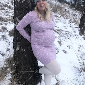 May include: A pregnant person wearing a light purple dress with a white pattern, white tights, and black boots. They are standing next to a birch tree in a snowy landscape.