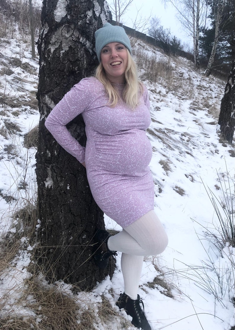 May include: A pregnant person wearing a light purple dress with a white pattern, white tights, and black boots. They are standing next to a birch tree in a snowy landscape.