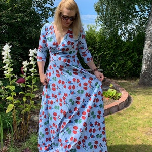 May include: A woman wearing a light blue maxi dress with a floral print of red flowers, green leaves, and blue birds. The dress has a V-neckline and 3/4 sleeves.