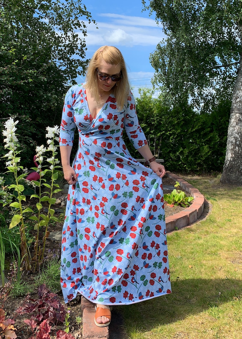 May include: A woman wearing a light blue maxi dress with a floral print of red flowers, green leaves, and blue birds. The dress has a V-neckline and 3/4 sleeves.
