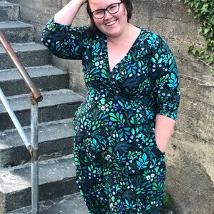 May include: A woman wearing a green and blue floral print wrap dress with 3/4 sleeves and pockets. She is standing on a set of concrete steps.