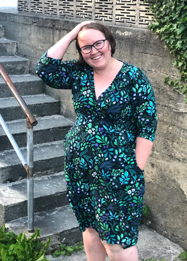 May include: A woman wearing a green and blue floral print wrap dress with 3/4 sleeves and pockets. She is standing on a set of concrete steps.
