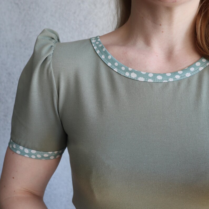 Puede incluir: Una camiseta verde salvia con ribete de lunares blancos alrededor del cuello y las mangas.