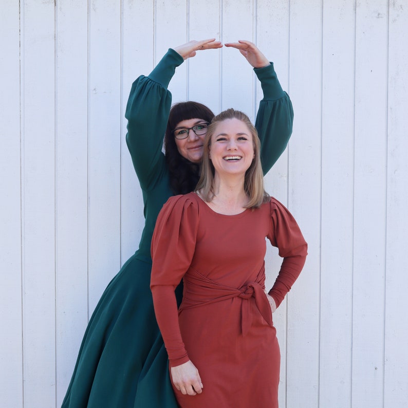 Puede incluir: Dos mujeres con vestidos se paran frente a una pared blanca. La mujer con el vestido verde est&aacute; sosteniendo sus brazos por encima de su cabeza, formando un c&iacute;rculo. La mujer con el vestido color &oacute;xido est&aacute; sonriendo y mirando a la c&aacute;mara.