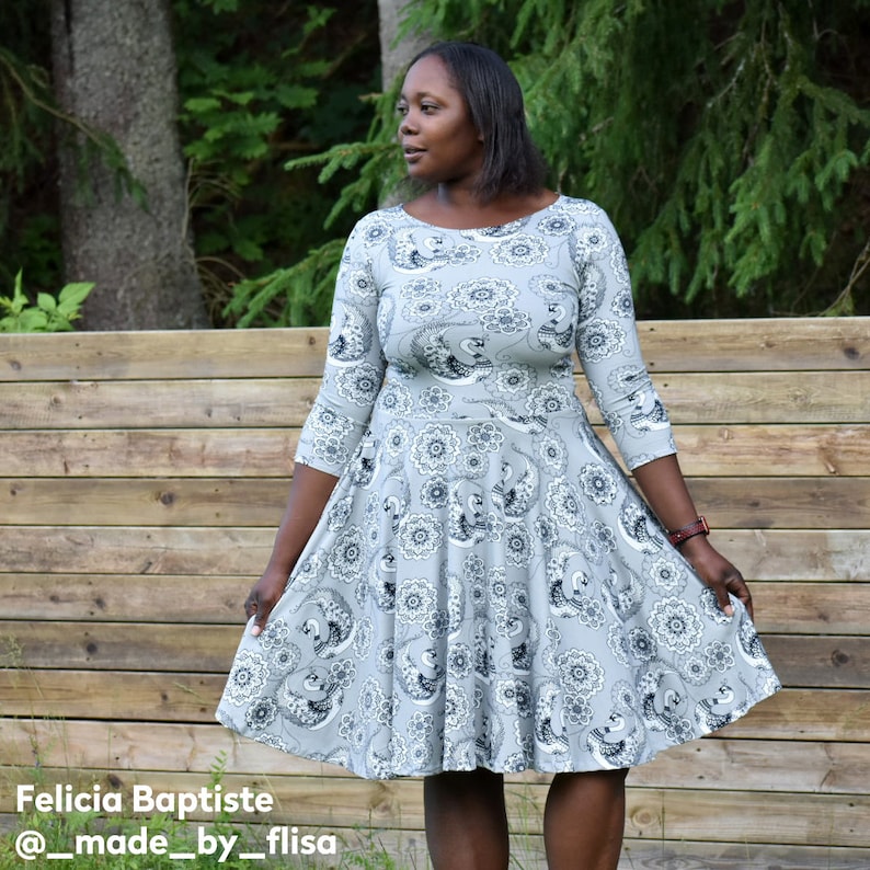 May include: A woman wearing a gray dress with a black and white floral pattern. The dress has a round neckline and 3/4 sleeves. The dress is fitted at the waist and flows out to a full skirt. The woman is standing in front of a wooden fence.