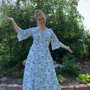May include: A woman wearing a long, white dress with a colorful floral and animal print. The dress has a V-neckline and 3/4 sleeves. She is standing in a garden with green foliage and brick pavers.