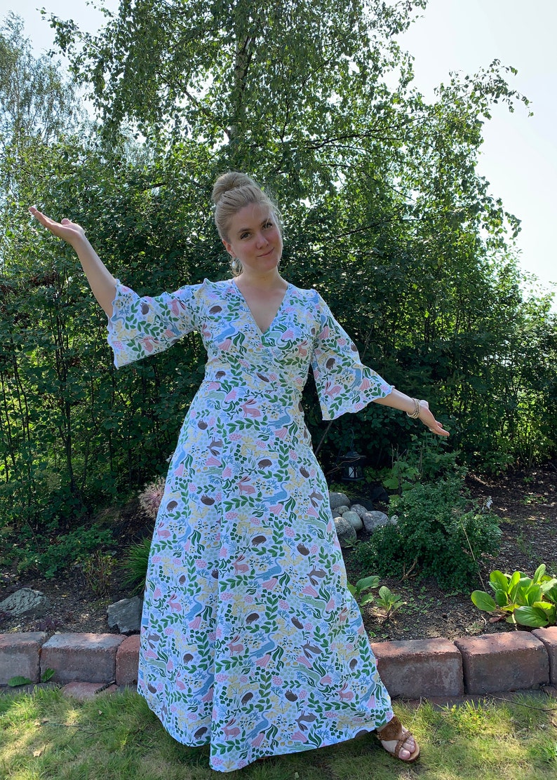 May include: A woman wearing a long, white dress with a colorful floral and animal print. The dress has a V-neckline and 3/4 sleeves. She is standing in a garden with green foliage and brick pavers.