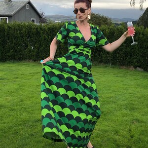 May include: A woman wearing a green and black maxi dress with a scale pattern, sunglasses, and earrings. She is holding a glass of pink champagne and standing in a grassy field.
