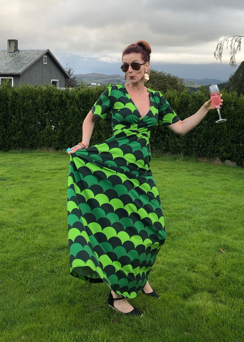 May include: A woman wearing a green and black maxi dress with a scale pattern, sunglasses, and earrings. She is holding a glass of pink champagne and standing in a grassy field.