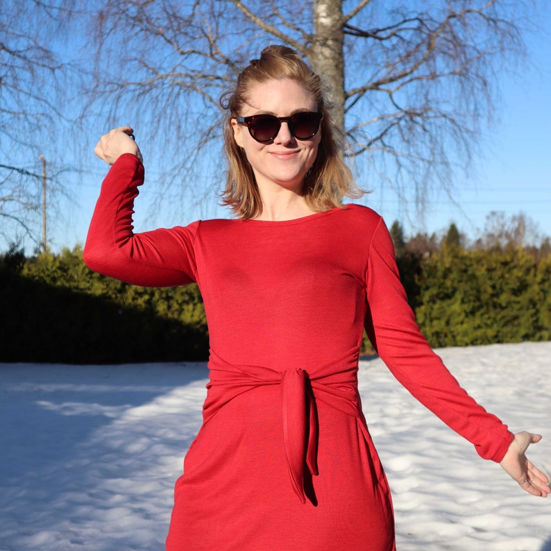 Puede incluir: Una mujer con un vestido rojo de manga larga con un lazo en la cintura. Lleva gafas de sol y tiene el pelo recogido hacia atr&aacute;s. El fondo es un paisaje nevado.