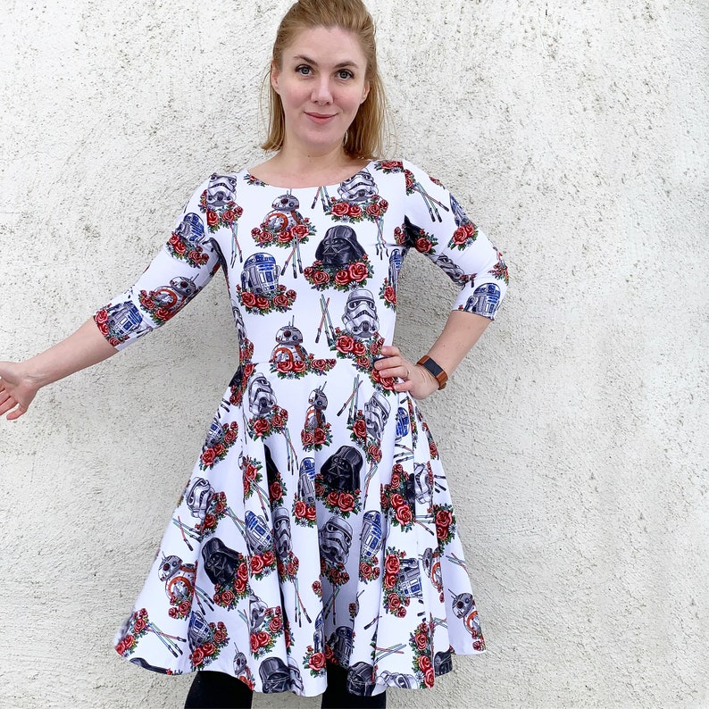May include: A white dress with a floral print featuring Star Wars characters, including Darth Vader, R2-D2, and C-3PO. The dress has a fitted bodice and a flared skirt.