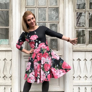 May include: A woman wearing a black dress with a pink floral print. The dress has a 3/4 sleeve and a flared skirt. She is standing in front of a white door with a window.