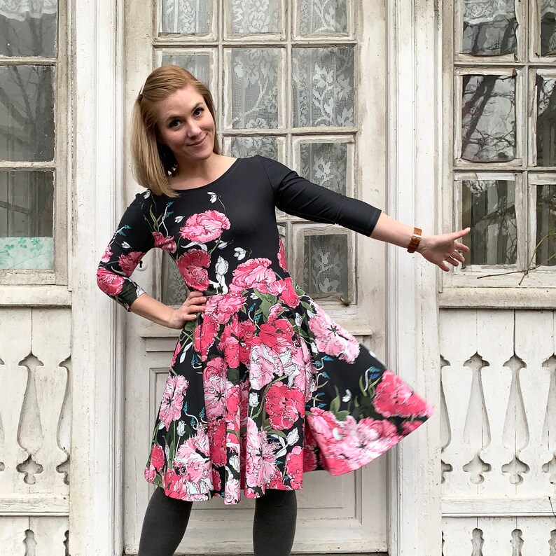 May include: A woman wearing a black dress with a pink floral print. The dress has a 3/4 sleeve and a flared skirt. She is standing in front of a white door with a window.