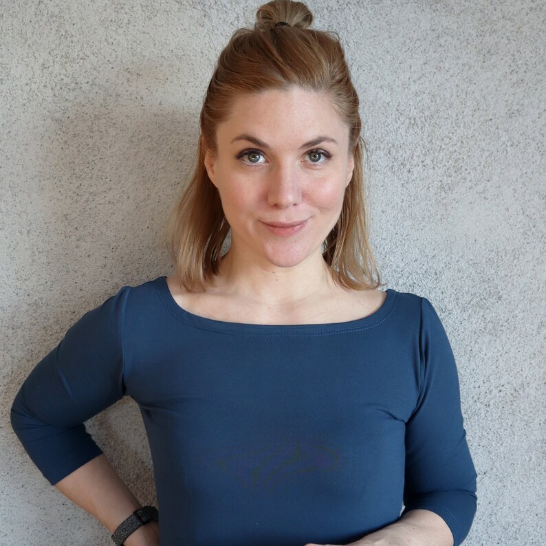 Puede incluir: Una mujer con el pelo largo y casta&ntilde;o claro lleva una camisa azul oscuro de manga larga con cuello redondo. Est&aacute; de pie frente a una pared gris texturizada.