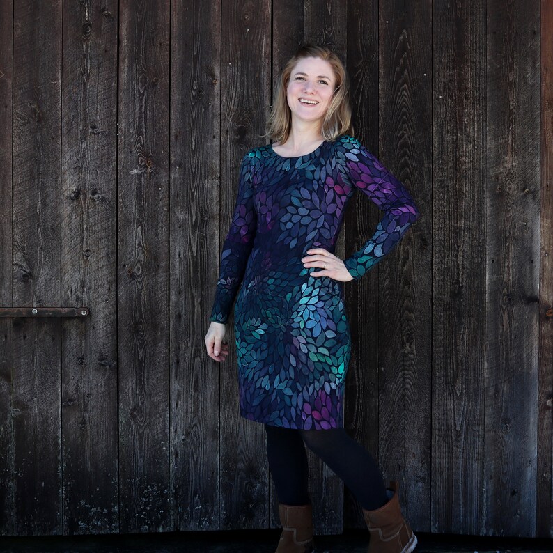 Puede incluir: Una mujer lleva un vestido de manga larga con un estampado abstracto de hojas en verde azulado, morado y negro. El vestido tiene un escote redondo y mangas largas. Est&aacute; de pie frente a una pared de madera.