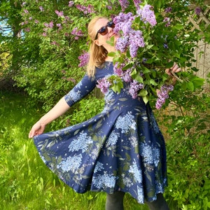 May include: A woman in a blue floral dress with a white background is standing in a garden. She is holding a bouquet of purple lilac flowers and smiling.