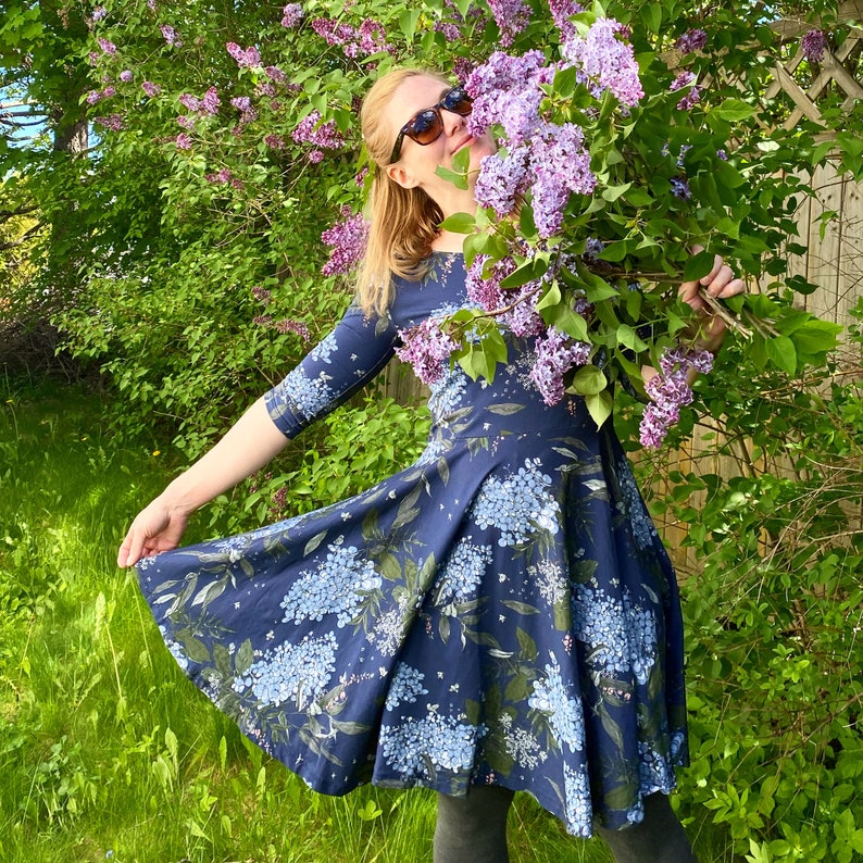 May include: A woman in a blue floral dress with a white background is standing in a garden. She is holding a bouquet of purple lilac flowers and smiling.