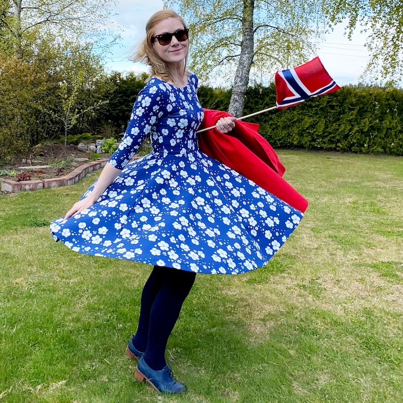 May include: A woman wearing a blue and white floral dress with a red jacket draped over her arm. She is holding a small Norwegian flag in her right hand.