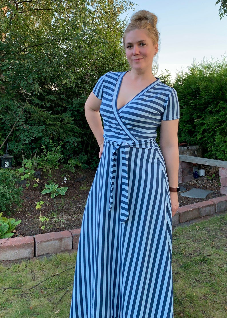 May include: A woman wearing a blue and white striped maxi dress with a wrap style bodice and a tie belt. The dress has short sleeves and a V-neckline.