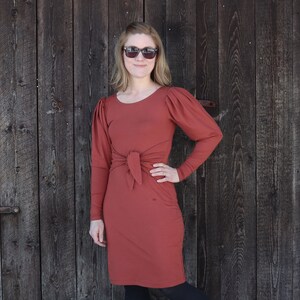 Puede incluir: Un vestido de color &oacute;xido con mangas largas y un lazo en la cintura. El vestido tiene un ligero detalle de manga abullonada. La modelo lleva gafas de sol.
