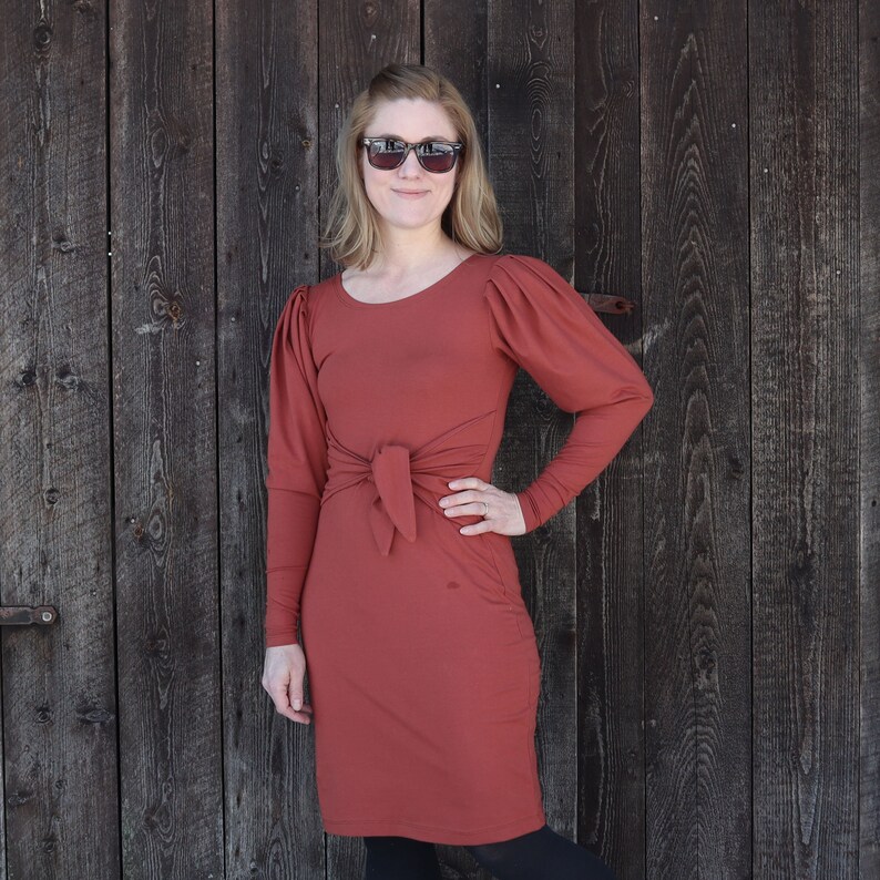 Puede incluir: Un vestido de color &oacute;xido con mangas largas y un lazo en la cintura. El vestido tiene un ligero detalle de manga abullonada. La modelo lleva gafas de sol.