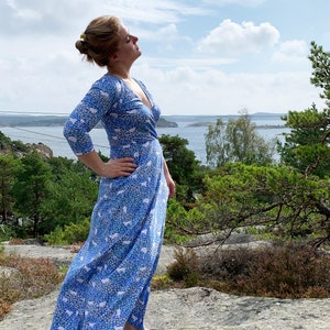 May include: A woman wearing a blue and white patterned maxi dress stands on a rocky surface with a view of a body of water and trees in the background.