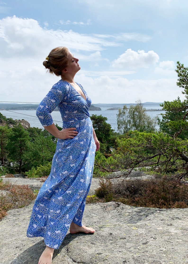 May include: A woman wearing a blue and white patterned maxi dress stands on a rocky surface with a view of a body of water and trees in the background.
