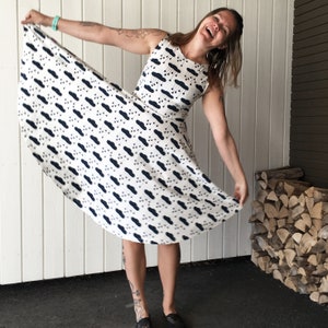 May include: A white dress with a black and white cloud and raindrop pattern. The dress has a sleeveless design and a flared skirt. The person wearing the dress is smiling and holding the skirt out.