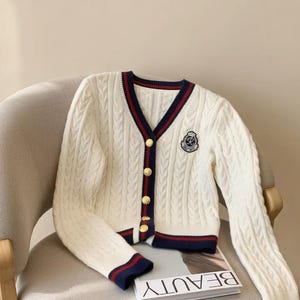 May include: Cream-colored cable knit cardigan with navy and red trim. The V-neck sweater features gold buttons and a crest emblem on the chest. The word "BEAUTY" is visible on a book beneath the cardigan. The cardigan is on a chair.