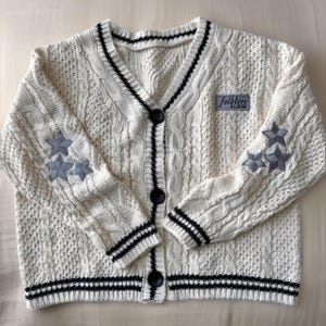 May include: Cream-colored cable knit cardigan with black trim and buttons. Features three gray star patches on each sleeve and a "folklore album" patch. The V-neck sweater has a classic design.
