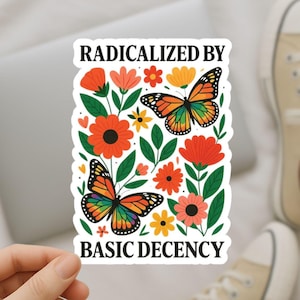 May include: A sticker with the text "RADICALIZED BY BASIC DECENCY" features two butterflies with rainbow wings and orange, red, and yellow flowers. The sticker is held by a hand, with a laptop and sneakers in the background.