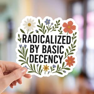 May include: A white sticker with the text "RADICALIZED BY BASIC DECENCY" surrounded by a floral border. The sticker is held by a hand against a blurred background. The flowers are in shades of white, orange, blue, and yellow.