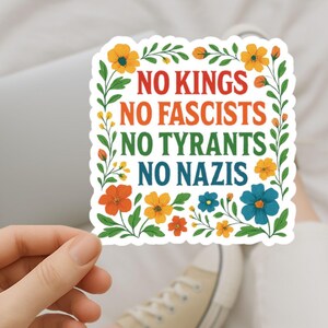 May include: A square sticker with the text "NO KINGS NO FASCISTS NO TYRANTS NO NAZIS" in red, orange, green, and blue. The text is surrounded by a floral border with orange, yellow, and blue flowers and green leaves.
