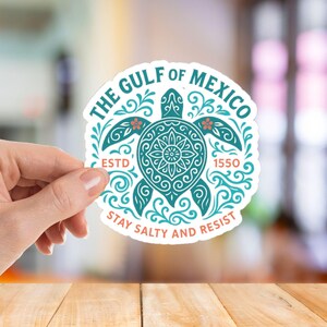 May include: A round sticker with a teal turtle design and the text "THE GULF OF MEXICO" and "ESTD 1550". The sticker also says "STAY SALTY AND RESIST" in red and white. The sticker is held by a hand.