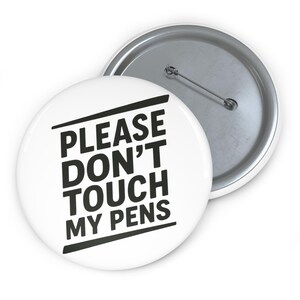 May include: Two white round buttons with a metal pin backing. The front button has the text "PLEASE DON'T TOUCH MY PENS" in bold black letters. The second button is partially visible, showing the metal backing.