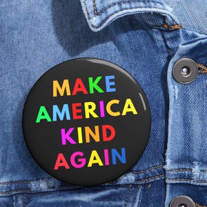 May include: A round, black button with the text "MAKE AMERICA KIND AGAIN" in rainbow colors. The button is pinned to a blue denim jacket, with the button's text prominently displayed.