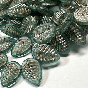 May include: A collection of teal-colored leaf-shaped beads with a metallic gold-colored vein design. The beads are made of glass and are scattered across a white surface. The beads are approximately 1 inch in length.