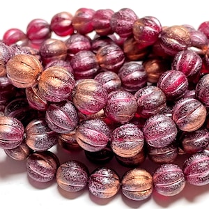 May include: Close-up of a collection of round beads in shades of burgundy, copper, and pink. The beads have a textured, ridged surface. The beads are strung together, suggesting they are for jewelry making or crafting.