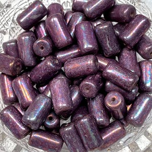 May include: A collection of dark purple, cylindrical beads with a glossy, iridescent finish. The beads are piled in a clear, textured glass bowl. Each bead has a hole running through the center, suitable for stringing.
