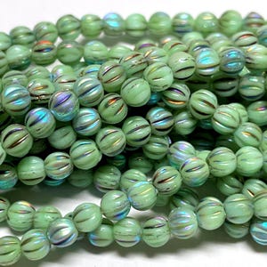 May include: A close-up of a strand of light green glass beads. Each bead is round with a ribbed design and iridescent accents. The beads are strung together, creating a textured and colorful display, perfect for jewelry making.