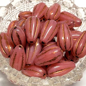 May include: A collection of pink, teardrop-shaped beads with gold stripes. The beads are clustered together in a clear, decorative glass bowl. The beads have a glossy finish and are ideal for jewelry making or crafting projects.