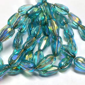 May include: A collection of translucent, teardrop-shaped glass beads in a vibrant turquoise blue hue. Each bead features a faceted surface with a shimmering, iridescent finish that reflects light, creating a multi-dimensional appearance.