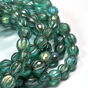 May include: Close-up of strands of teal-colored beads. The beads have a faceted, oval shape with a metallic silver accent. The beads are strung together, creating a layered effect. The beads have an iridescent quality, reflecting light.