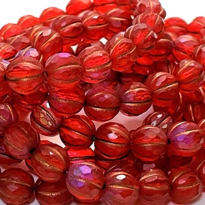 May include: A collection of faceted, round beads in a deep red hue. Each bead features gold-colored stripes and a textured surface, creating a striking visual effect. The beads are strung together, suggesting their use in jewelry making or crafting.