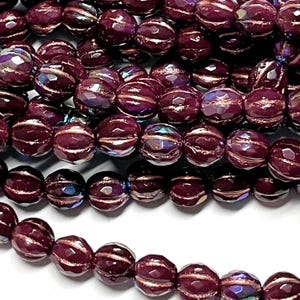 May include: Close-up of a collection of faceted, round beads in a deep burgundy color. Each bead has a metallic gold stripe around the center. The beads have an iridescent sheen, reflecting multiple colors.