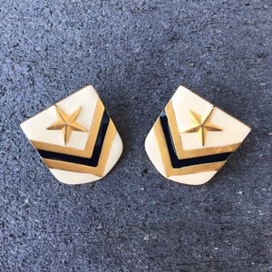 May include: A pair of gold and black chevron earrings with a gold star in the center. The earrings are on a gray background.