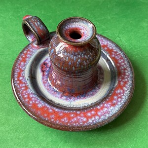 May include: A ceramic candle holder with a red, white, and brown glaze. The holder has a round base and a tall, cylindrical candle cup with a handle.