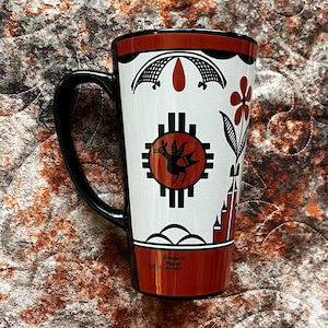 May include: Tall ceramic mug with a black handle and rim, featuring a white body adorned with Native American-inspired designs in black and red. The mug includes a floral motif and a circular emblem with a bird. The text "Elizabeth Medina" is printed on the mug.